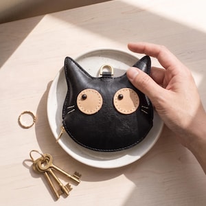Könnte beinhalten: Eine schwarze Ledergeldbörse in Form eines Katzenkopfes mit hellbraunen, kreisförmigen Augenaufnähern, kleinen dunklen Pupillen und gestickten Schnurrhaaren. Das Etui hat einen goldfarbenen D-Ring und einen goldenen Reißverschluss.