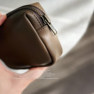 Könnte beinhalten: Eine kleine, braune Leder-Reißverschlusstasche mit einem Metall-Reißverschluss-Zipper. Die Tasche ist geschlossen und der Reißverschluss ist nach oben gezogen.