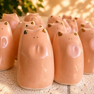 Puede incluir: Una colección de pequeñas figuras de cerdo de cerámica color melocotón. Cada figura presenta un acabado liso y brillante, dos pequeñas orejas pintadas de dorado, dos puntos diminutos para los ojos y una hendidura circular como hocico.