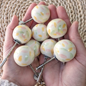 Puede incluir: Una colección de ocho pomos de cerámica con una base blanca y un patrón colorido de amarillo, verde y rosa. Los pomos tienen un vástago de metal plateado.