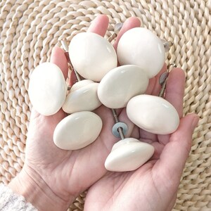 Peut inclure: Un ensemble de boutons de cabinet en céramique blanche avec des ferrures argentées. Les boutons sont disposés dans une main, mettant en valeur leur forme ronde et lisse.