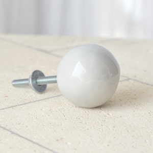 Puede incluir: Pomo de cerámica blanco con un tornillo plateado. El pomo es redondo y tiene un acabado liso y brillante.