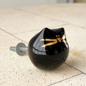 Pode incluir: Botão de cerâmica preto com bigodes e nariz pintados a ouro. O botão tem um parafuso prateado.