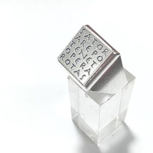 Puede incluir: Un anillo de plata con el cuadrado de palabras latinas "SATOR AREPO TENET OPERA ROTAS" grabado en la parte superior. El anillo se exhibe sobre un soporte transparente y geométrico sobre un fondo blanco. El diseño del anillo es simple y elegante.