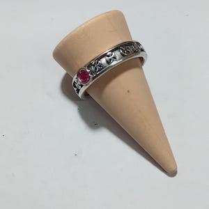 Op de afbeelding: Een zilveren ring met een decoratief patroon en een kleine, ronde, rode edelsteen. De ring wordt getoond op een lichtbruine, kegelvormige standaard tegen een witte achtergrond. Het ontwerp van de ring heeft herhaalde bloem- of kruisachtige vormen.