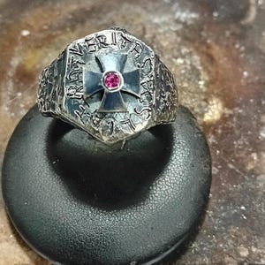 May include: A silver ring with a black cross and a red stone in the center. The ring is engraved with the words "VERITAS" and "FRATRES".