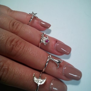 Puede incluir: Una mano con cuatro anillos de plata con diferentes colgantes. Los colgantes incluyen una estrella de mar, una luna creciente, un pájaro pequeño y un corazón con una piedra rosa.