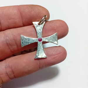 Könnte beinhalten: Ein silberner Kreuzanhänger mit gehämmertem Muster und einem kleinen, runden, roten Edelstein in der Mitte. Das Kreuz hängt an einer feinen Silberkette. Der Anhänger wird in einer Hand vor weißem Hintergrund gehalten.