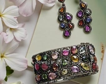 May include: A pair of drop earrings and a wide hinged cuff bracelet, both crafted from dark metal and encrusted with multi-colored, pear-shaped gemstones in shades of pink, yellow, green, and blue. The jewelry set features the text Jewels Fine Hub.