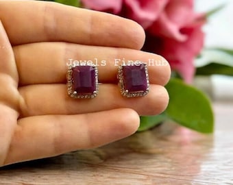 Pode incluir: Um par de brincos retangulares com pedras centrais de cor magenta profundo rodeadas por um halo de pequenas pedras claras cravadas numa armação metálica. O texto Jewels Fine Hub é visível no centro da imagem.