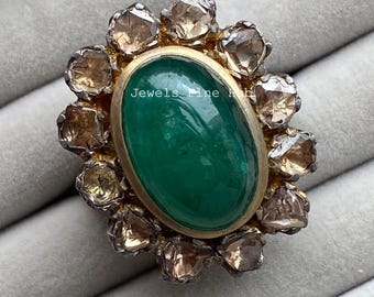 May include: An oval emerald ring with a gold setting, encircled by a halo of light brown gemstones. The ring is displayed on a grey surface. The text "Jewels Fine Hub" is visible.