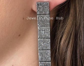 May include: A long, rectangular earring with a series of stacked, square sections. Each section is covered in small, sparkling stones, creating a shimmering effect. The earring is silver-toned and appears to be worn on an ear. The text "Jewels Fine Hub" is visible.