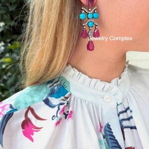 Puede incluir: Un pendiente tipo candelabro con engaste de metal oscuro, piedras turquesas redondas, piedras magenta en forma de lágrima y pequeños cristales transparentes. El texto Jewelry Complex es visible en el lado derecho del diseño.