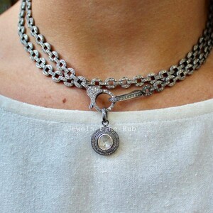 Op de afbeelding: Zilverkleurige ketting met een dubbel kettingontwerp, met een ronde hanger met een centrale steen. De ketting en hanger zijn versierd met kleine, sprankelende details. De ketting heeft een sluiting.