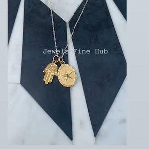 May include: A silver chain necklace with two gold pendants. One pendant is a Hamsa hand, and the other is an oval compass design. The text "Jewels Fine Hub" is visible. The background is a black and white geometric pattern.