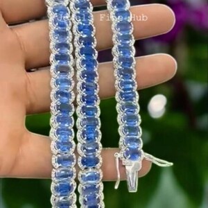 May include: A silver-toned tennis bracelet featuring a continuous row of oval-cut blue gemstones, each surrounded by a halo of small clear stones. The metal clasp is visible at the end. The text Jewels Fine Hub is printed on the bracelet.