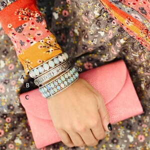 May include: Three silver bracelets with opal stones are stacked on a wrist. The bracelets are worn with a pink clutch. The person is wearing a floral patterned top with orange and yellow accents.
