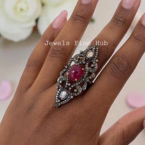 May include: An elongated, dark metal ring featuring a central cabochon-cut ruby surrounded by intricate filigree metalwork and small, clear, rose-cut diamonds. The text Jewels Fine Hub is visible on the surface of the ring setting.