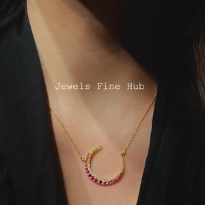 May include: A gold-colored crescent moon pendant necklace. The moon is encrusted with red and clear gemstones. The necklace is worn on a person with a black top. The text "Jewels Fine Hub" is visible.