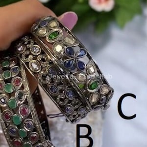 Puede incluir: Tres brazaletes de metal plateado etiquetados A, B y C. El brazalete A presenta piedras ovaladas rojas y verdes. Los brazaletes B y C presentan piedras azules, verdes y transparentes en patrones florales. El texto Jewels Fine Hub es visible en el metal.
