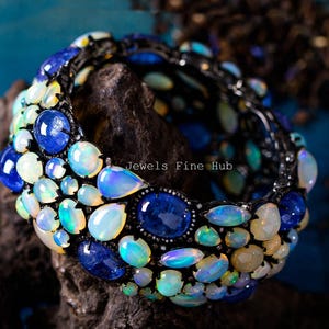 May include: A striking bracelet featuring a variety of gemstones. The bracelet is adorned with blue sapphires and opals in various shapes and sizes. The text "Jewels Fine Hub" is visible on the image.