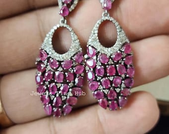 Pode incluir: Brincos pendurados prateados com um conjunto de pedras preciosas rosa-escuras de corte oval. Os brincos apresentam um design em forma de gota, com um halo de pequenas pedras transparentes. O texto "Jewels Fine Hub" é visível.