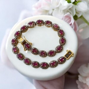 Könnte beinhalten: Ein goldfarbenes Armband mit einer Reihe ovaler roter Edelsteine, die jeweils von einem Kranz aus kleinen klaren Steinen umgeben sind. Das Schmuckstück verfügt über einen rechteckigen Verschluss mit dekorativem Muster und Hakenmechanismus.