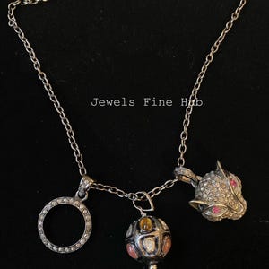 Puede incluir: Tres collares de plata con diferentes colgantes. Un colgante es un círculo, otro una esfera con piedras de colores y el tercero una cabeza de gato con ojos rosas. El texto "Jewels Fine Hub" es visible.
