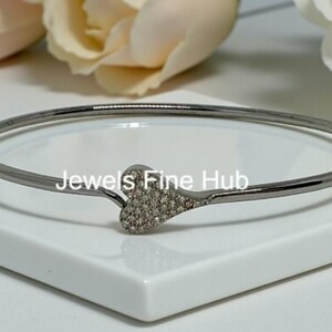 Könnte beinhalten: Ein dunkelmetallischer Armreif mit einem herzförmigen Anhänger, der mit kleinen klaren Steinen besetzt ist. Der Text "Jewels Fine Hub" ist in der Mitte des Armreifs aufgedruckt. Der Armreif hat eine glatte, polierte Oberfläche und ein dünnes Band.