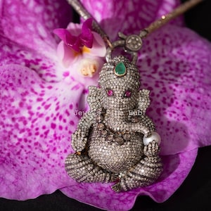 Peut inclure: Un collier pendentif détaillé représentant une figure de Ganesh assise, incrustée de nombreux petits diamants. Le pendentif a des yeux en rubis et un accent émeraude. Le collier repose sur une fleur d'orchidée violette.