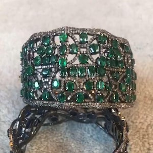 Puede incluir: Una pulsera ancha y ornamentada con una montura de metal oscuro, incrustada con numerosas piedras preciosas de color verde esmeralda. La pulsera presenta un dise&ntilde;o calado y est&aacute; adornada con piedras m&aacute;s peque&ntilde;as y transparentes. El texto "Jewels Fine Hub" es visible.