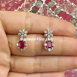 May include: A pair of gold-toned earrings featuring a floral cluster design at the top and a rectangular, deep red gemstone at the center. The gemstones are surrounded by smaller, clear stones. The earrings are displayed on a light-toned background.