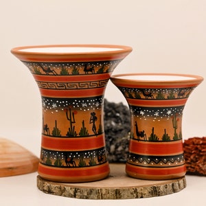 May include: Two terracotta ceramic bowls with a white interior and painted designs. The bowls feature a geometric pattern, a cactus, and a llama. The bowls are on a wooden base.