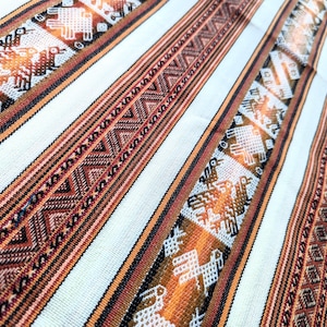 Op de afbeelding: Close-up van een witte stof met geweven bruine, oranje en zwarte strepen. De strepen bevatten geometrische patronen en gestileerde dierfiguren.