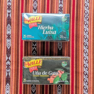 May include: Two boxes of herbal tea bags. The top box is blue and green and says "Hierba Luisa" with a picture of the herb. The bottom box is black and green and says "U&ntilde;a de Gato" with a picture of the herb.