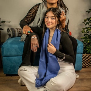 May include: Two women wearing scarves, one in a black and white animal print top and the other in a black top with a blue scarf. They are sitting on a blue couch with a patterned throw pillow.