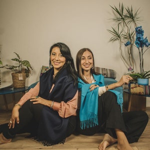 May include: Two women are sitting on a wooden floor, wearing blue and turquoise scarves. The woman on the left is wearing a pink shirt and the woman on the right is wearing a black shirt. They are both smiling at the camera.