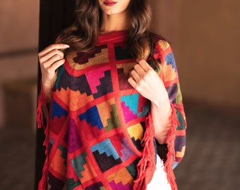 Può includere: Un poncho colorato con un motivo geometrico nei toni del rosso, arancione, blu e verde. Il poncho ha un orlo con frange ed è realizzato in tessuto a trama. L'indumento è indossato sopra pantaloni bianchi.