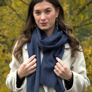 May include: A woman wearing a navy blue scarf and a light beige coat. The scarf is draped around the neck and has a fringed edge. The background features a blurred view of yellow and green foliage.