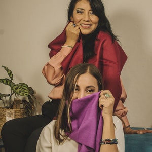 May include: Two women wearing scarves, one in a burgundy scarf and the other in a purple scarf. The woman in the burgundy scarf is smiling and holding the scarf up to her face. The woman in the purple scarf is covering her face with the scarf.