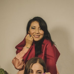 May include: A woman with long black hair is smiling and wearing a burgundy scarf. She is wearing a pink shirt and a ring on her finger.