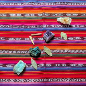 Op de afbeelding: Een assortiment kristallen, gedroogde bladeren en Palo Santo-stokken is gerangschikt op een kleurrijk, gestreept textiel. De kristallen omvatten amethist, citrien en turkooizen stenen. Het textiel heeft een levendige mix van rode, oranje, roze en blauwe strepen.