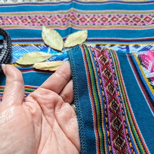 May include: Close-up of a blue fabric with colorful stripes and intricate patterns. The fabric is folded over a hand, showing the detail of the stitching.