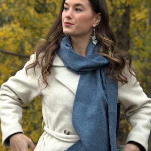 May include: A woman wearing a cream-colored coat and a blue scarf. The scarf is draped around the neck. She is also wearing silver heart-shaped earrings. The background features fall foliage.