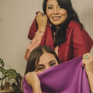 May include: Two women wearing scarves. The woman in the foreground is wearing a purple scarf and is covering her face with it. The woman in the background is wearing a red scarf and is holding it up to her face.