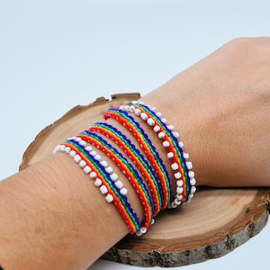 Könnte beinhalten: Ein Regenbogen-Freundschaftsarmband mit weißen Perlen. Das Armband ist aus geflochtener Schnur gefertigt und wird um ein Handgelenk gebunden.