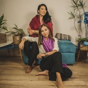 May include: Two people are sitting on a blue couch. The person on the left is wearing a red scarf and the person on the right is wearing a purple scarf. They are both looking at the camera.
