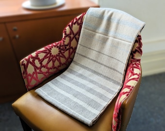 Puede incluir: Una manta de tiro a rayas en tonos beige y gris está colocada sobre un sillón marrón con tapicería de terciopelo rojo. El sillón tiene un patrón geométrico en el respaldo.