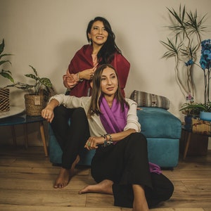 May include: Two women sitting on a blue couch, one wearing a burgundy shawl and the other wearing a purple scarf. They are both looking at the camera and smiling.