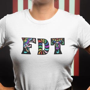 Peut inclure: T-shirt blanc avec les lettres "FDT" dans une police décorative colorée. La personne porte un pantalon de camouflage et se tient devant un drapeau américain.